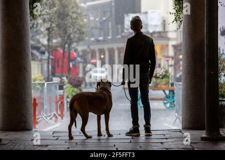 Un uomo e il suo cane si affacciano su una piazza deserta di Covent Garden durante una doccia a pioggia primaverile, nel centro di Londra, in Inghilterra, nel Regno Unito Foto Stock