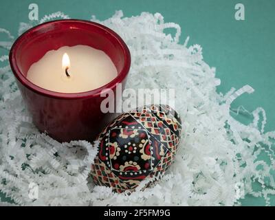 Uovo di Pasqua colorato e candela bruciante su carta bianca grattugiata. Tradizione pasquale Foto Stock