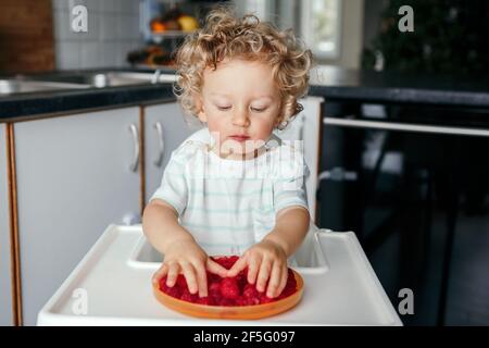 Mangiare sano. Carino bambino caucasico che mangia frutta rossa matura a casa. Divertente bambino seduto seggiolone con frutti di bosco freschi in cucina. Supplemento Foto Stock