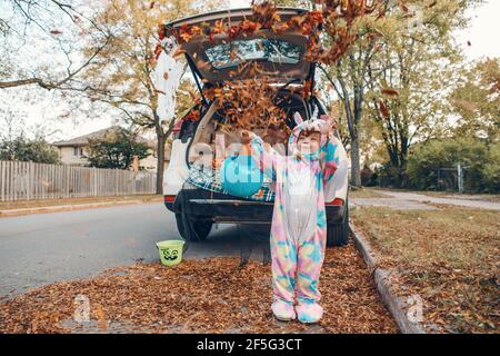 Trucco o tronco. Bambino felice in costume unicorno che lancia foglie e celebra Halloween in tronco di auto. Carino bambino sorridente che si prepara per ottobre ho Foto Stock