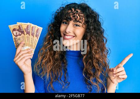 Giovane ragazza ispanica che tiene 5000 banconote forint ungheresi sorridenti felici puntare con la mano e il dito verso il lato Foto Stock