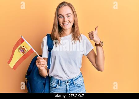 Bella donna bionda scambio studente tenendo bandiera spagnola sorridente felice e positivo, pollice su facendo eccellente e segno di approvazione Foto Stock