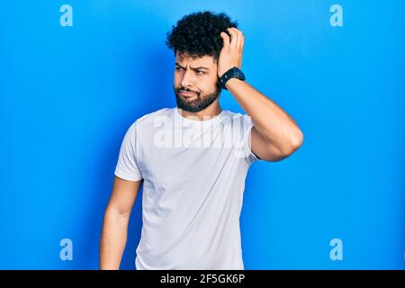Il giovane arabo con la barba che indossa una t-shirt bianca casual confondono e si chiedono domande. Incerto con il dubbio, pensando con la mano sulla testa. Pensivo Foto Stock