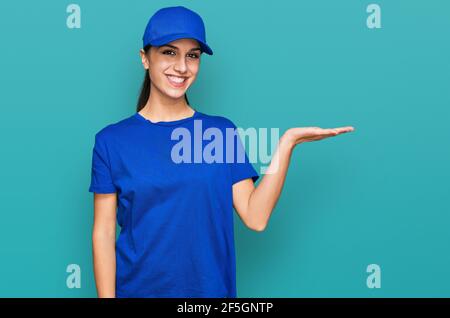 Giovane ragazza ispanica che indossa uniforme corriere di consegna sorridente allegro presentazione e puntando con palmo di mano guardando la fotocamera. Foto Stock