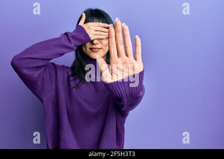 Giovane donna bruna con pangs che indossa un pullover turtleneck che copre gli occhi con le mani e facendo il gesto di stop con espressione triste e paura. Imbarazzato a. Foto Stock