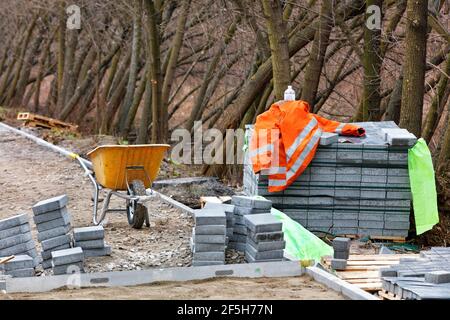 Una giacca arancione del costruttore si trova su una pila di lastre di pavimentazione vicino a una carriola gialla sullo sfondo di alberi in crescita nel parco. Foto Stock