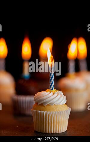 Primo piano immagine vista laterale di un assortimento di cupcake al cioccolato e alla vaniglia con glassature bianche e marroni. Ognuno ha una candela accesa contro bac scuro Foto Stock