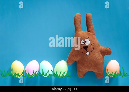 Caccia di Pasqua con coniglietto giocattolo e uova colorate in vetro su sfondo blu Foto Stock