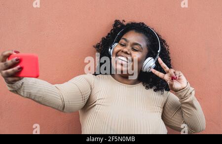 Felice donna africana che prende selfie con smartphone mobile durante l'ascolto musica con le cuffie wireless Foto Stock