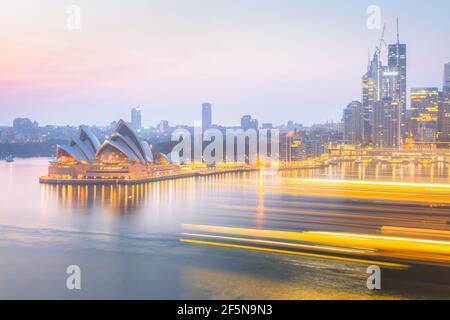 Vista del paesaggio urbano all'alba dal Ponte dell'Harbour sullo skyline del Porto di Sydney e il teatro dell'opera nel New South Wales, Australia, mentre una grande barca passa lascia dietro una luce Foto Stock