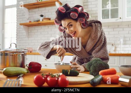 Donna divertente casalinga in arricciacapelli e accappatoio in piedi cottura fresco piatto sano Foto Stock