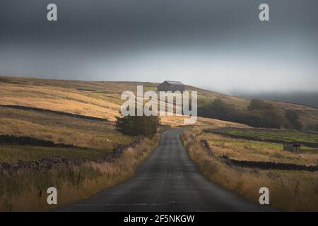 Un vecchio fienile di pietra e una strada di campagna vuota attraverso una luna campagna inglese paesaggio rurale nel Nord Pennines AONB, Inghilterra Regno Unito. Foto Stock