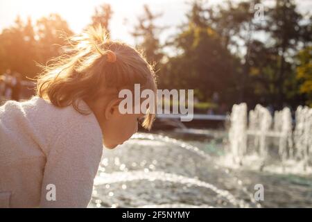 adorabile bambina guarda il laghetto con fontane nel parco in una giornata di sole. weekend a piedi in famiglia. passare il tempo con i bambini Foto Stock