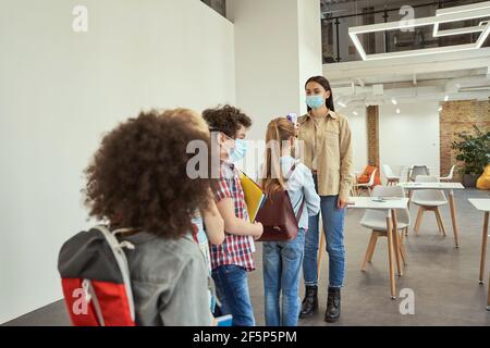 Una giovane insegnante studia i bambini della scuola per la febbre contro la diffusione di Covid19, usando un termometro digitale mentre i bambini vengono a scuola Foto Stock