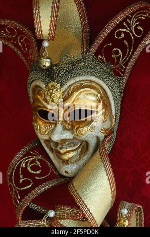 Una tipica maschera carnevalesca veneziana raffigurante Arlecchino disegnata in stile tradizionale stile con foglia d'oro e finitura della vernice incrinata su a. sfondo rosso Foto Stock