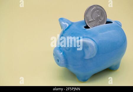 Banco di porcellini in ceramica blu con un aspetto sospetto e una moneta di 5 CZK nel foro su sfondo giallo. Spazio di copia Foto Stock