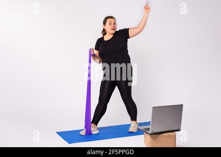 Attività domestica e formazione, corso di fitness online. Donna bionda sovrappeso che fa esercizi di stretching, banda di resistenza violetta che guarda video guida Foto Stock