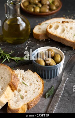 Pane italiano alla ciabatta a fette su trivello con erbe, olio extra vergine e olive marinate su sfondo scuro grunge Foto Stock