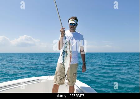 Pescatore della Florida che tiene un piccolo Tunny (Euthynnus alletteratus) sospeso da un gancio di pula mentre è in piedi nella prua Di una barca con l'Atlantico Oce Foto Stock