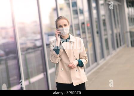Donna sicura che indossa la maschera facciale e parla con qualcuno sul telefono cellulare mentre cammina di fronte al business center durante la pandemia del coronavirus. Foto Stock