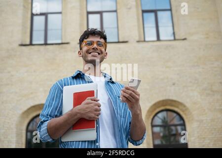 Ritratto di giovane bello studente indiano che tiene portatile, libro, telefono cellulare, a piedi per l'università. Sorridente uomo d'affari asiatico utilizzando lo smartphone Foto Stock