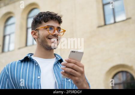 Un uomo indiano che parla sul cellulare, cammina per strada, sorridendo. Ritratto di un ragazzo di stile che indossa abiti e occhiali casual utilizzando uno smartphone Foto Stock