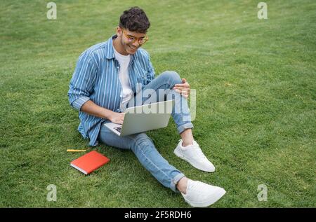 Felice studente asiatico che studia, impara la lingua, educazione online, seduto sull'erba. Giovane bell'uomo indiano che usa un computer portatile, che lavora all'aperto Foto Stock