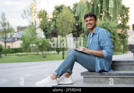 Programmatore indiano sorridente che usa il computer portatile, lavoro freelance progetto online, seduto in parco. Studio di studenti asiatici, concetto di istruzione Foto Stock