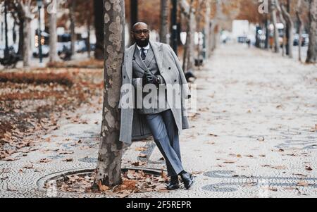 Un cappero nero imprenditore senior in occhiali e un elegante vestito con un costume su misura e un cappotto, si appoggia contro un albero su un autunno b Foto Stock