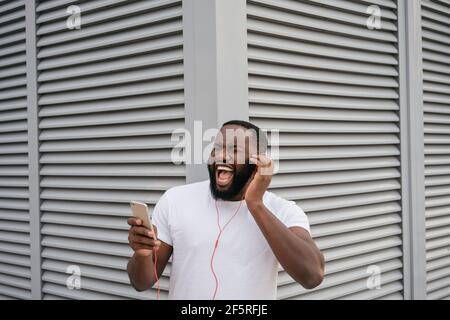 Impressionabile afroamericano che ascolta musica all'aperto, canti canti. Hipster felice indossando abbigliamento casual danzare su strada urbana, divertendosi Foto Stock