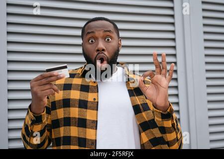 Eccitato afroamericano uomo tenendo carta di credito per lo shopping online, mostrando segno OH Foto Stock