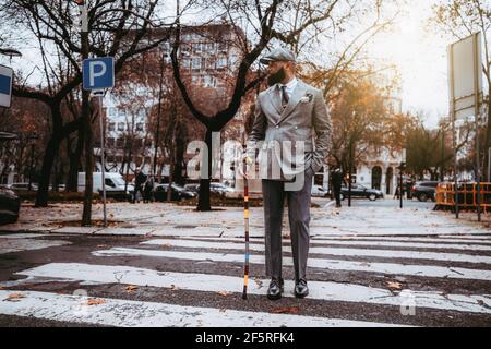 Un cappero nero sopportato imprenditore senior in un elegante costume su misura, cappello, con un bastone da passeggio colorato fantasia sta guardando da parte durante l'attraversamento Foto Stock