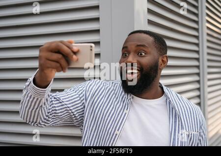 Eccitato afroamericano uomo prendendo selfie utilizzando il telefono cellulare in piedi su strada urbana. Video di ripresa emozionale dell'influencer blogger Foto Stock