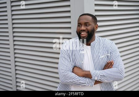 Primo piano ritratto di giovane confidente afro-americano con le braccia incrociate ridere, in piedi per strada. Business di successo Foto Stock