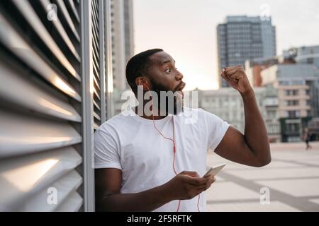 Un ragazzo hipster felice che indossa abiti casual danzando sulla strada urbana. Impressionabile afroamericano uomo che tiene il telefono mobile ascoltare la musica all'aperto Foto Stock