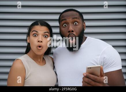 Eccitato afroamericano uomo e donna che tiene il telefono cellulare, shopping online guardando la macchina fotografica. Concetto di scommesse sportive Foto Stock