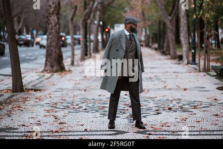 Un cappero africano anziano maturo con una barba nera ben curata è in piedi nel mezzo di un viale d'autunno e guardando da parte, con un elegante coa Foto Stock