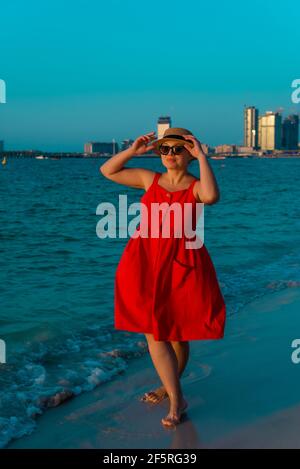 donna in abito rosso che cammina lungo la spiaggia ammirando il tramonto visualizza Foto Stock