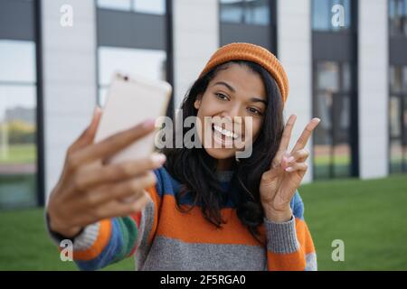 Giovane influencer emozionale blogger utilizzando il telefono cellulare, video in streaming online. Sorridente afroamericana che prende selfie, mostrando il segno della vittoria Foto Stock