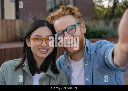 Giovani amici sorridenti che indossano occhiali eleganti prendendo selfie sulla strada. Bella donna asiatica e bell'uomo caucasico guardando la macchina fotografica Foto Stock