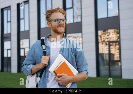 Studente universitario sorridente con zaino e libro a piedi nel campus universitario, concetto di istruzione. Ritratto di bell'uomo che indossa occhiali eleganti Foto Stock