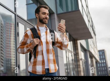 Uomo bearded usando il telefono mobile, in attesa del taxi all'aperto. Turista sorridente con zaino che tiene smartphone, cercando il modo migliore con la mappa di navigazione Foto Stock