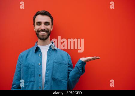 Ritratto di un giovane uomo sorridente che indossa una camicia casual che punta con la mano, isolato su sfondo rosso. Spazio di copia, concetto di pubblicità Foto Stock