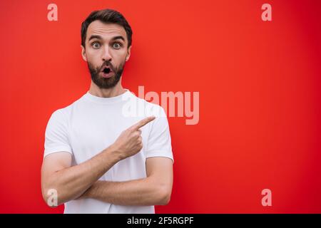 Ritratto di giovane uomo emozionale che indossa una t-shirt casual puntando con il dito, isolato su sfondo rosso. Spazio di copia, concetto di pubblicità Foto Stock
