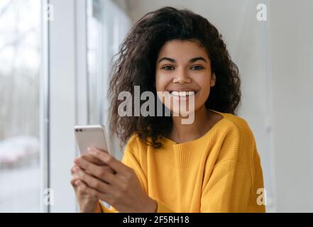 Bella donna afroamericana utilizzando il telefono cellulare, shopping online. Sorridente hipster che tiene smartphone, ordinare cibo, guardando la fotocamera Foto Stock