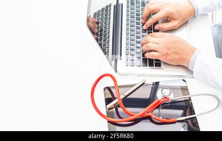Vista dall'alto delle mani di un medico che scrive sulla tastiera di un computer portatile accanto a un tablet e uno stetoscopio rosso su una scrivania bianca con un buon spazio per la copia. Medi Foto Stock
