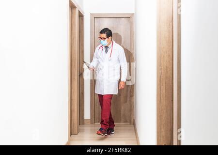 Medico ispanico con un cappotto bianco, cravatta, maschera e stetoscopio, visitando i suoi pazienti in un corridoio ospedaliero che tiene una tavoletta tra le mani. Salute worke Foto Stock