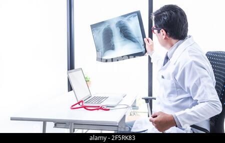 Medico che esamina una radiografia del torace e dei polmoni contro la luce della finestra del suo ufficio con un laptop e uno stetoscopio sulla sua scrivania. Concetto di medicina Foto Stock