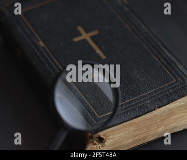 Chiusa Bibbia antica in pelle nera copertina con croce d'oro al centro e pagine giallastre è visto attraverso la lente di ingrandimento su sfondo nero. Ricerca Foto Stock