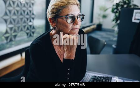 Donna d'affari anziana premurosa con la mano sul mento in ufficio. Donna professionista di affari che guarda via e pensa. Foto Stock